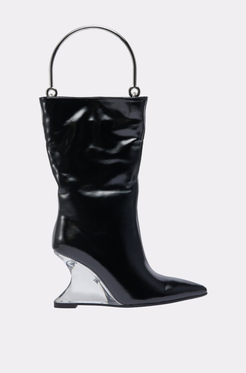 The STEP ON IT PURSE is a black, shiny bag shaped like a pointed-toe boot, featuring a silver metal handle and a transparent wedge-shaped heel, shown on a plain light background.
