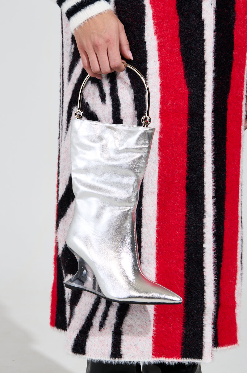 Clad in a red, black, and white striped outfit, a person holds the STANDIN ON BUSINESS BOOT PURSE. This eye-catching accessory is made from metallic leather, featuring a circular handle at the top of its boot design and displaying a bold novelty wedge heel shape.