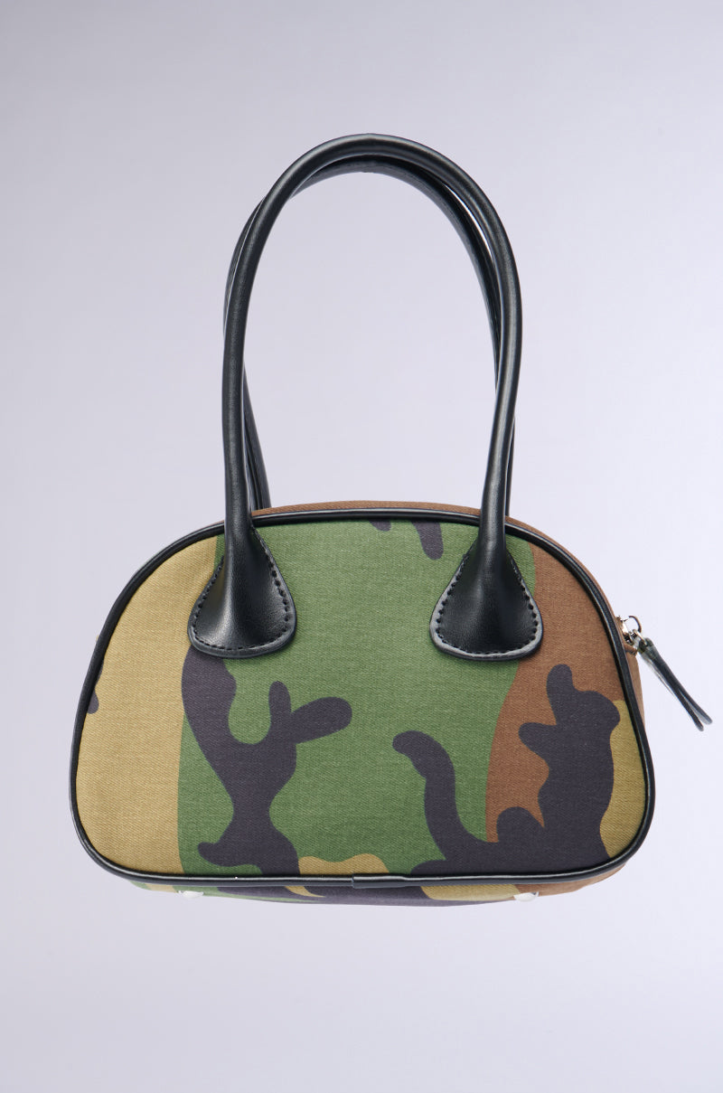 The KELLY CAMO NIGHTS MINI PURSE is a structured bowling bag that showcases a camouflage pattern in shades of green, brown, and black. It features sleek black leather handles and a metallic zipper closure, all set against a plain, light gray background.