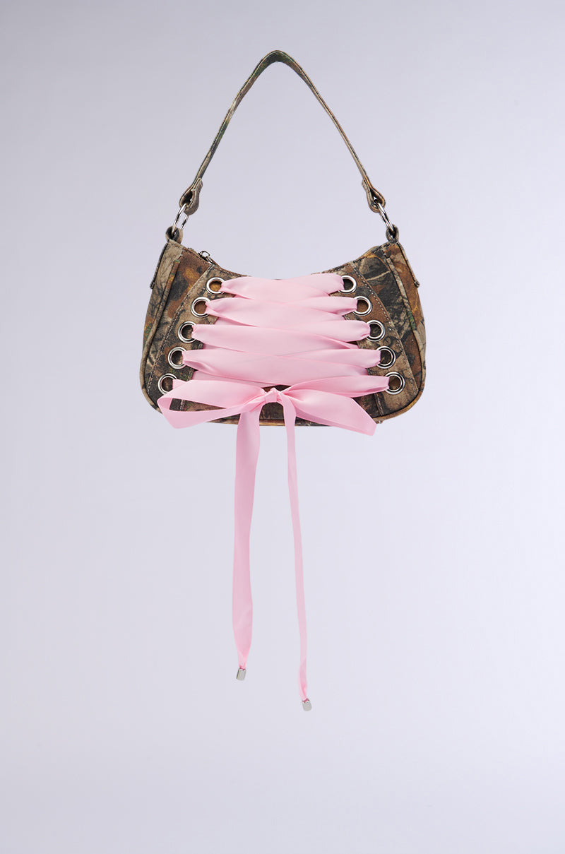 The MAKE IT FASHUN CAMO PURSE, a stylish fashion accessory with a brown, textured exterior, is adorned with large silver hardware grommets and pink ribbon laces crisscrossing over the front. This curved-shaped camo purse also features a single shoulder strap, all set against a light grey background.
