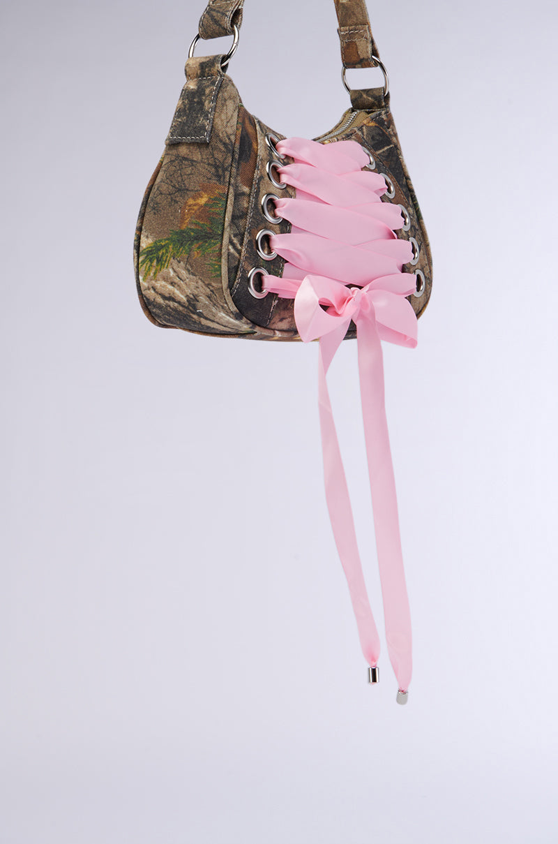 The MAKE IT FASHUN CAMO PURSE is a compact accessory adorned with a pink ribbon lace-up detail on the front, threaded through silver hardware eyelets and tied into a bow at the bottom. This stylish purse is showcased against a plain white background.