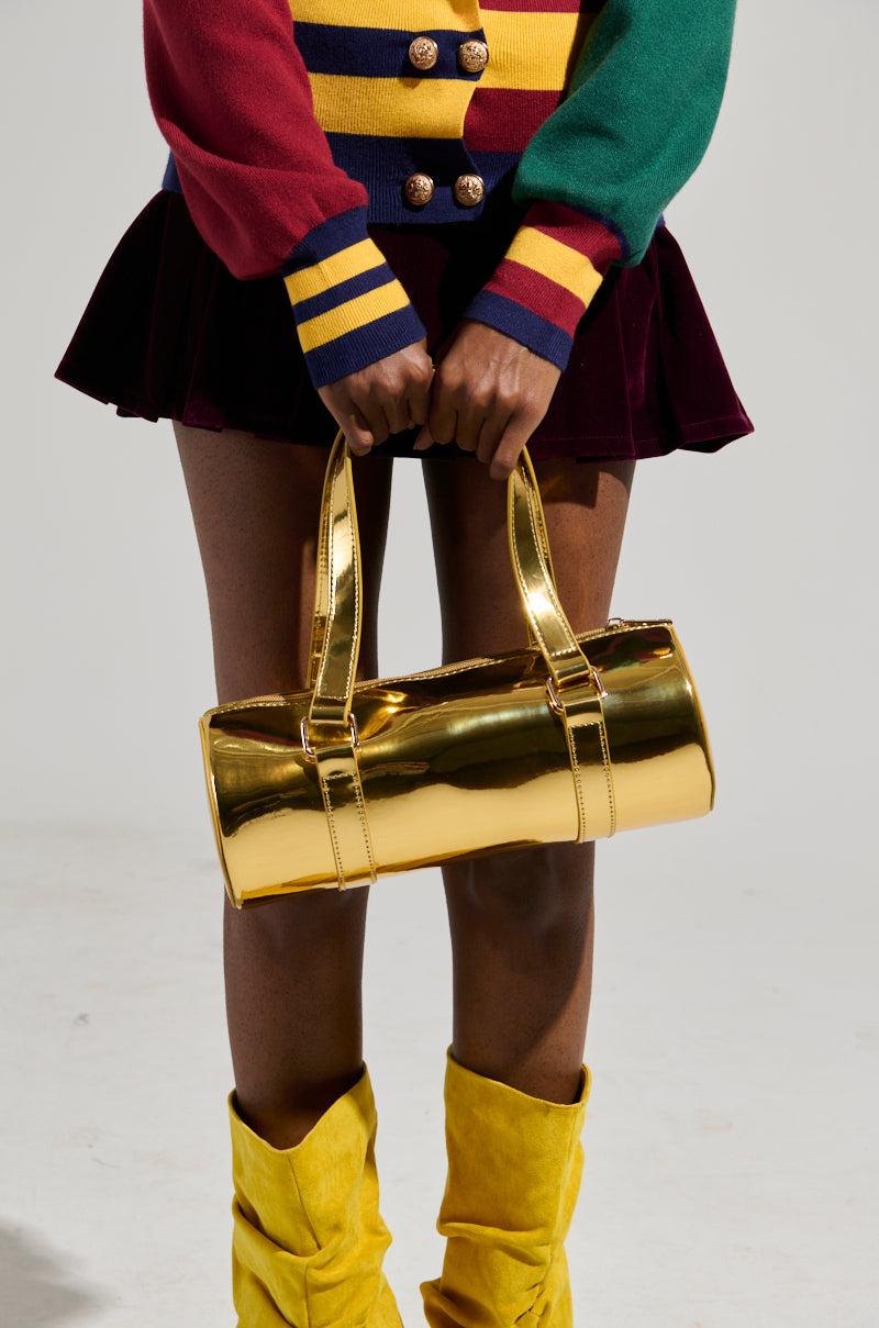 A person holds the CHROME DREAMIN PURSE while wearing a colorful striped top adorned with gold buttons, a dark skirt, and vibrant yellow boots. The image centers on the lower half of the ensemble, showcasing the striking accessory and fashion choices with its vivid barrel-style silhouette.