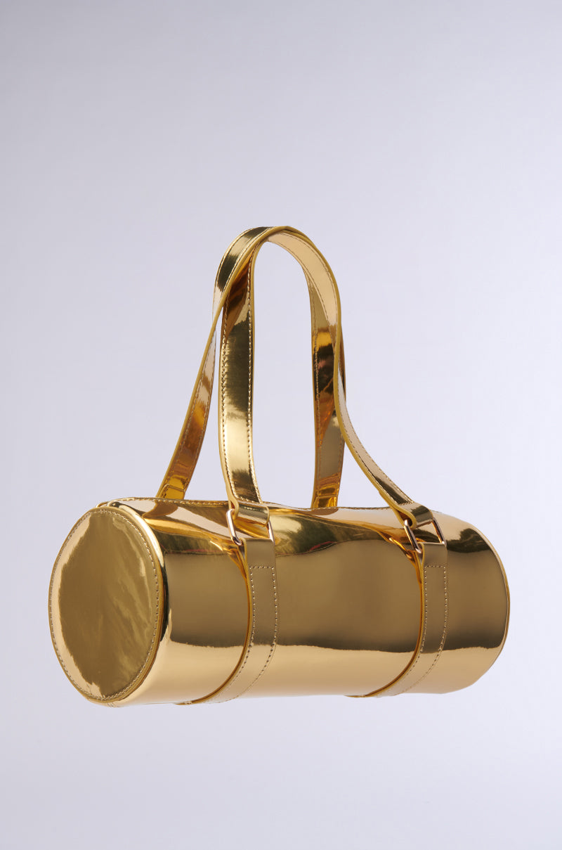 The CHROME DREAMIN PURSE, a lustrous gold handbag with a cylindrical barrel-style design and two coordinating straps, is crafted from faux leather and set against a neutral backdrop.