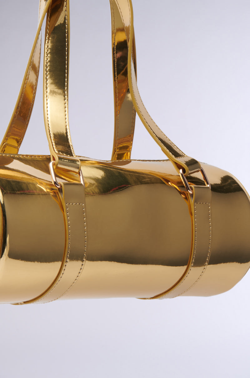 The CHROME DREAMIN PURSE, a shiny gold cylindrical handbag with matching gold straps, boasts a glossy faux leather finish and metallic appearance that reflects light beautifully. Suspended against a light gray background, its barrel-style silhouette creates a striking statement.