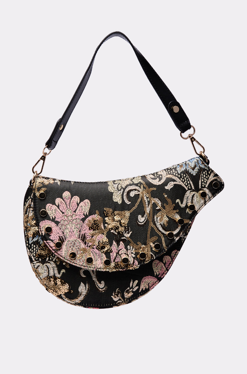 The BROCADE DREAMS SADDLE BAG is a crescent-shaped black brocade piece with a wide strap, gold, pink, and silver floral embroidery, decorative eyelets on the flap closure, and metallic accents for a sophisticated touch.