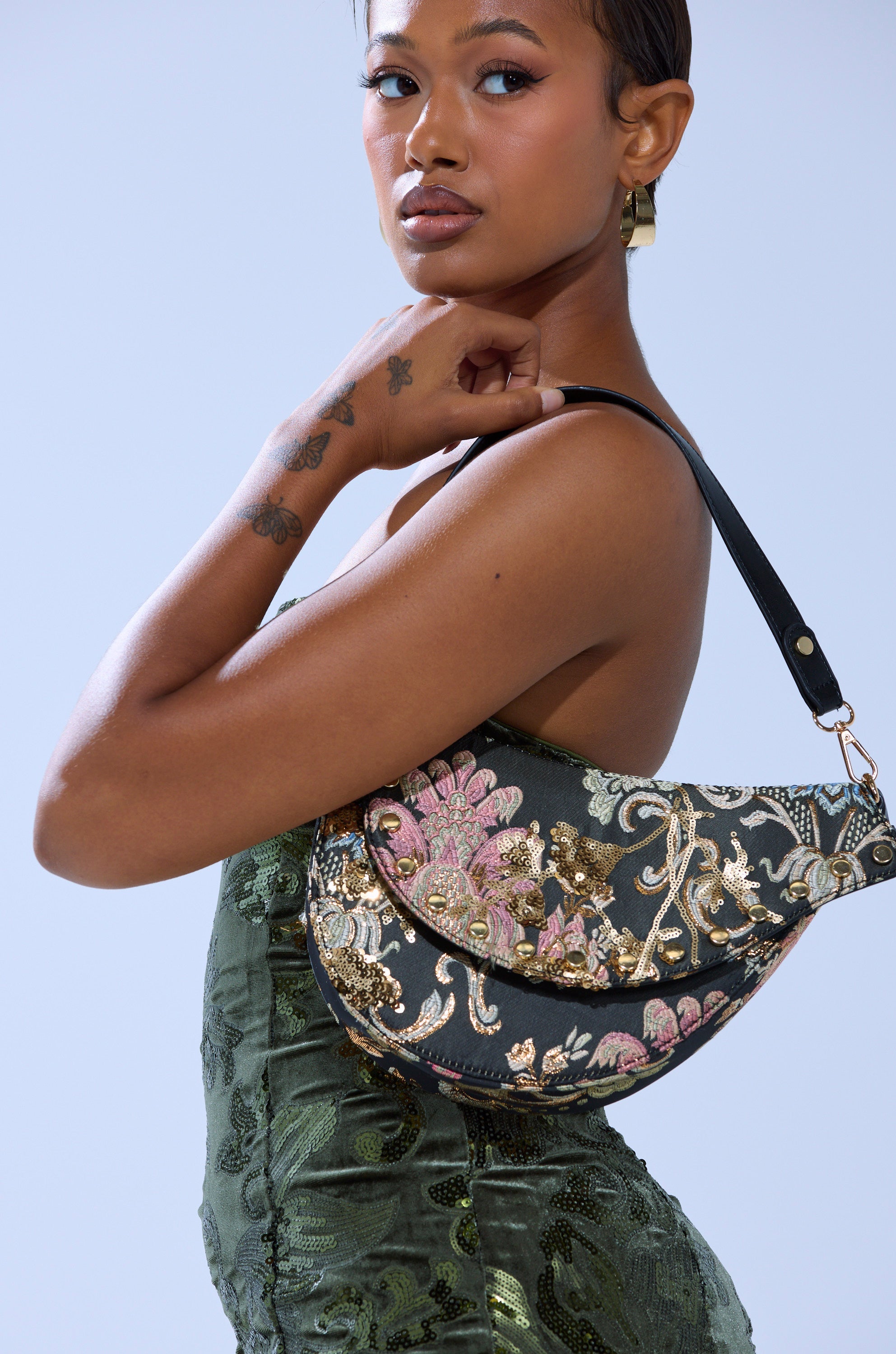A woman with short hair and visible forearm tattoos poses with the BROCADE DREAMS SADDLE BAG, featuring a structured saddle shape and gold hoop earrings. The bag is adorned with gold and pink floral embroidery. 1