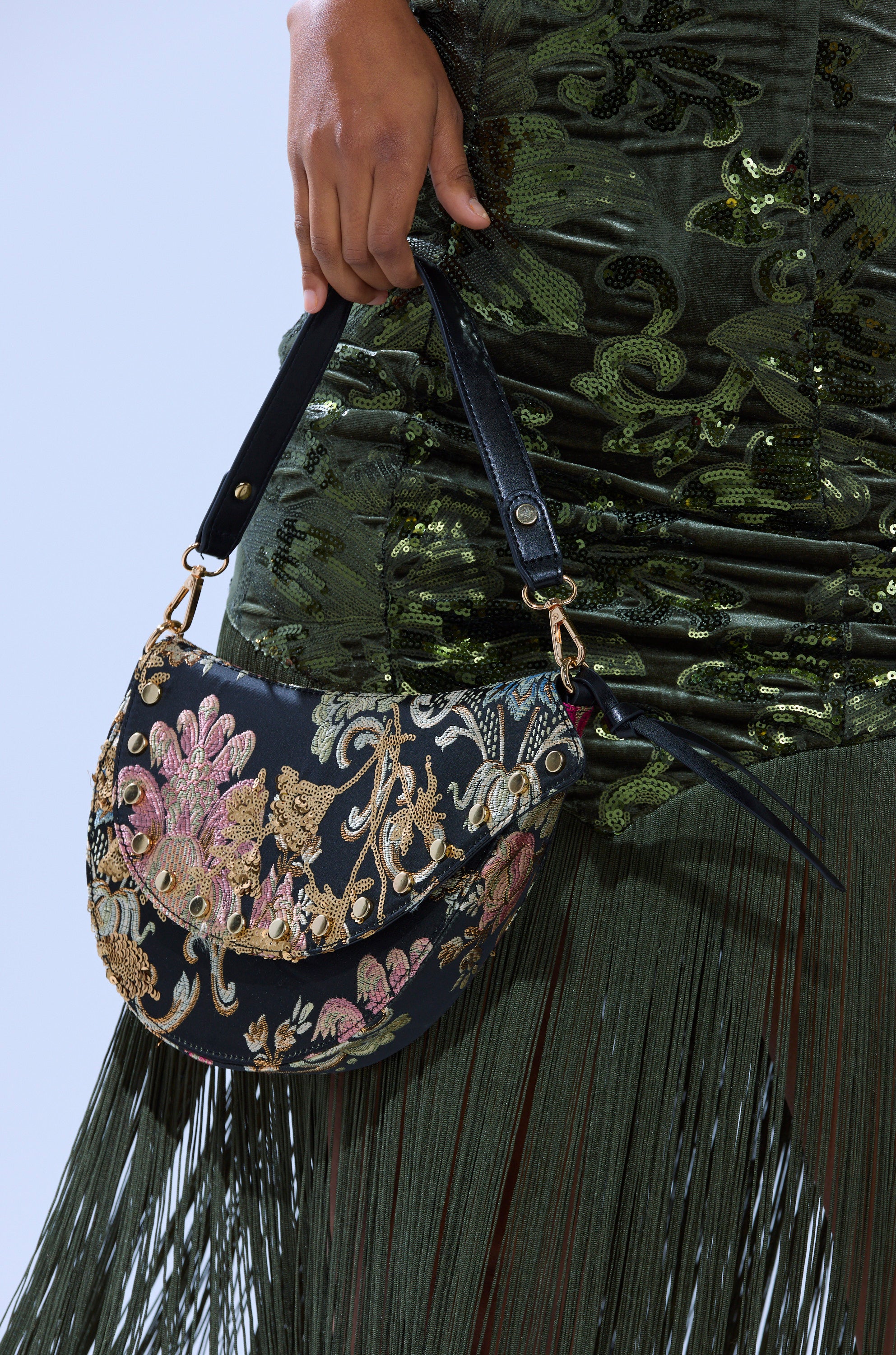 A person in a green sequin fringe dress holds the BROCADE DREAMS SADDLE BAG, a black brocade saddle bag adorned with intricate gold, pink, and blue floral embroidery.