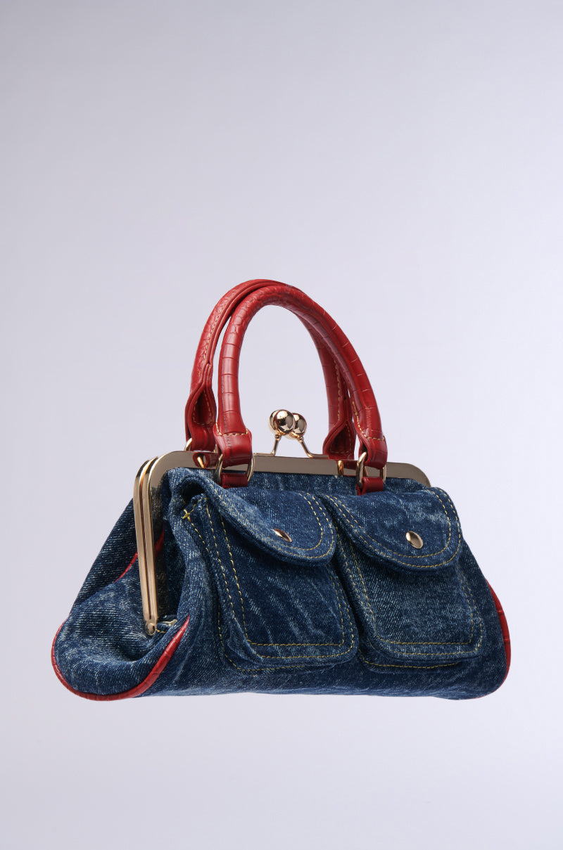 The ON THE FLIGHT CARGO COIN PURSE showcases a stylish and structured design with its denim exterior, complemented by red faux crocodile leather handles and accents. It features two front pockets and gold-tone hardware, including a clasp closure, all set against a neutral background.