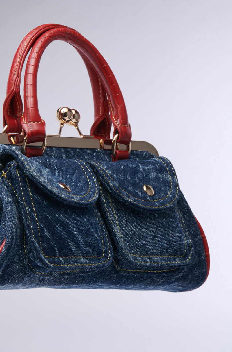 The ON THE FLIGHT CARGO COIN PURSE is a chic accessory crafted from acid wash denim, featuring two front pockets and shiny red leather handles and trim. Its design is completed with a metal clasp closure, all set against a neutral light gray background that highlights its elegant style.