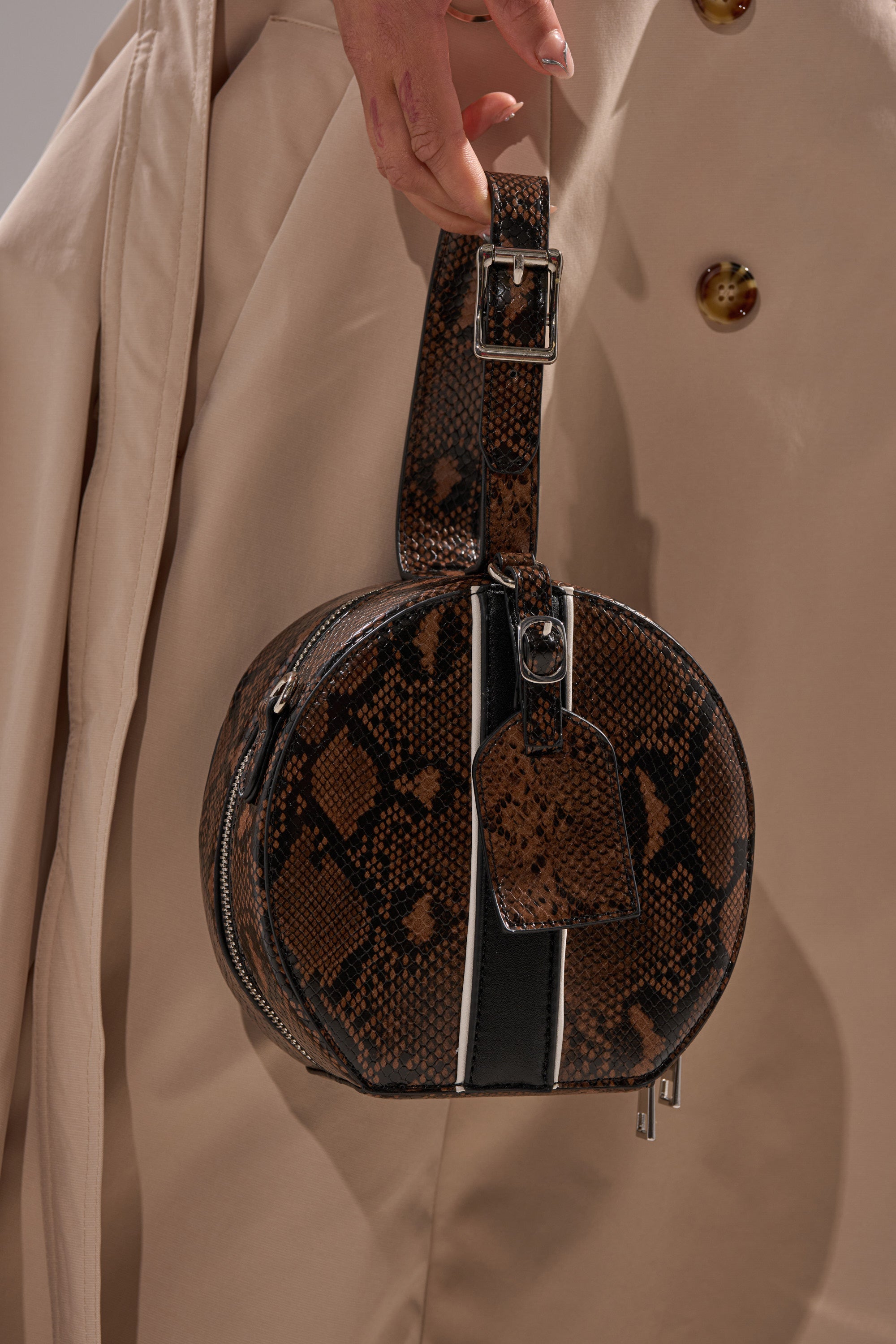A person in a beige coat holds the RENEGADE FAUX SNAKE PURSE, featuring embossed snakeskin, a black and white stripe, matching strap, silver buckle, zipper, and a removable crossbody strap. 1