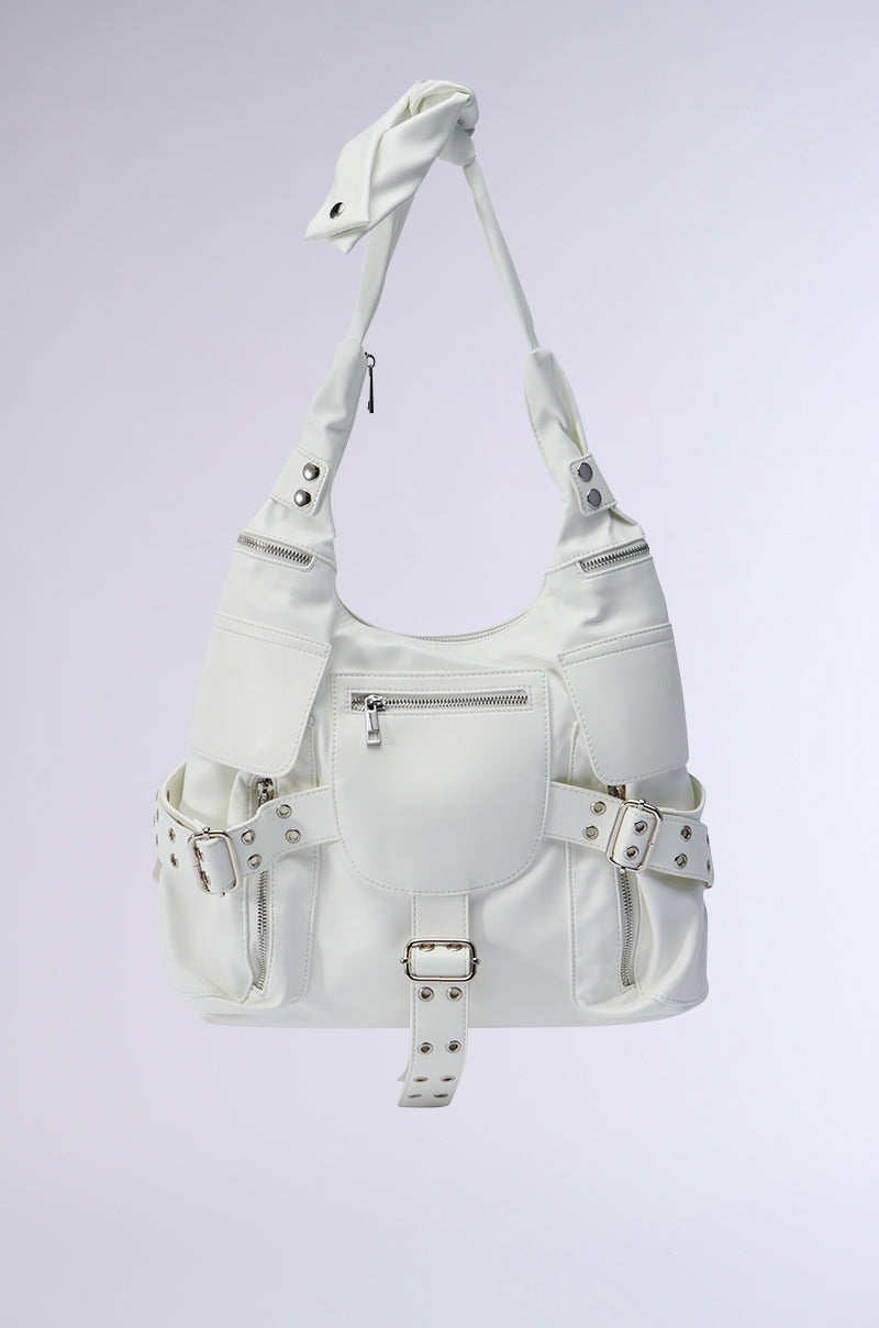 The R U WITH IT BAG is a stylish, roomy tote crafted in white faux leather. It showcases multiple compartments, silver hardware, and buckle accents. Its oversized design features a knotted strap and several studded exterior pockets set against a simple background. 0