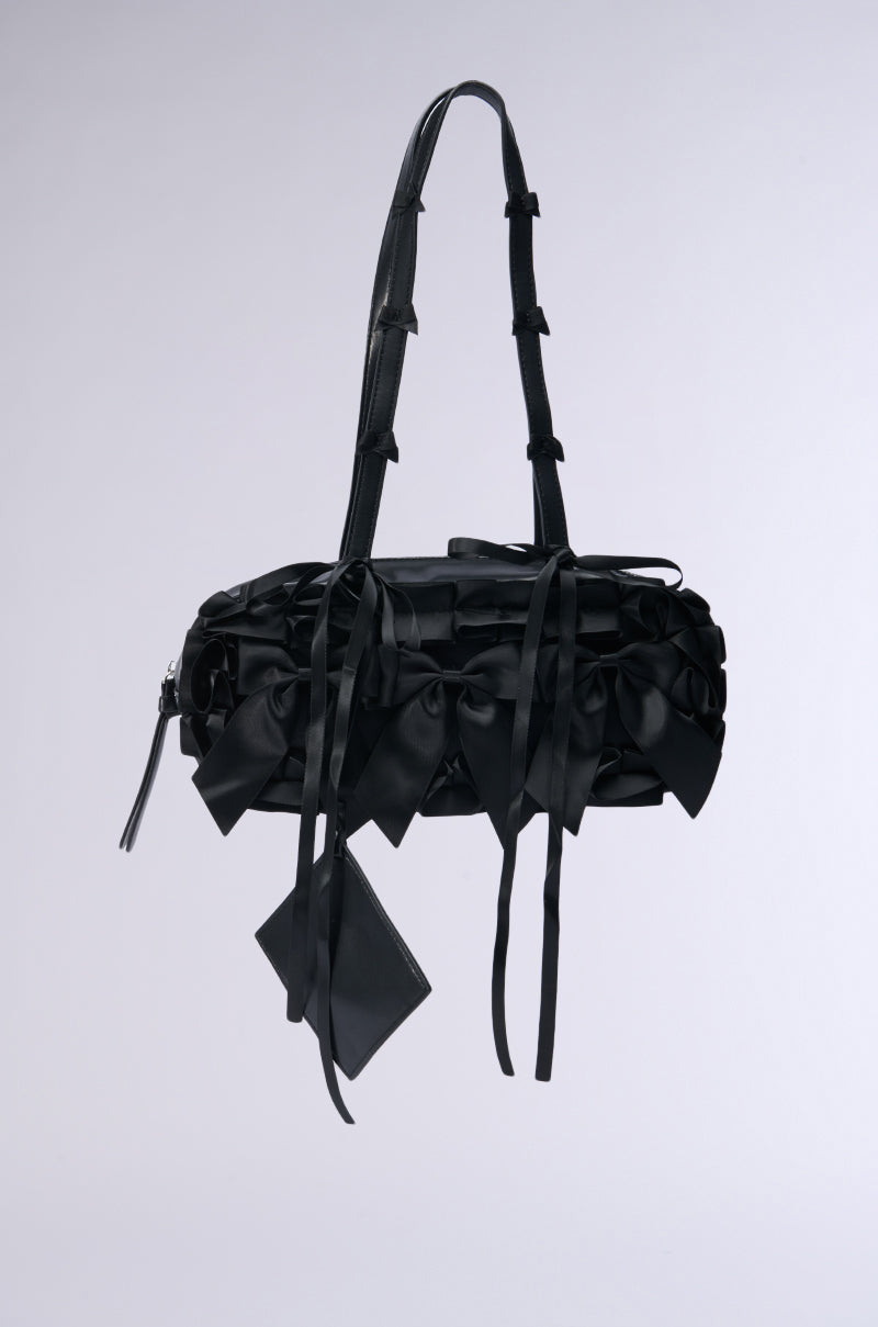 The BOWS GALORE PURSE is a chic black handbag embellished with numerous bows and ribbons on its faux leather surface. It includes a long, decorative strap and a small attached pouch, making this fashionable accessory stand out gracefully against a simple light gray background.