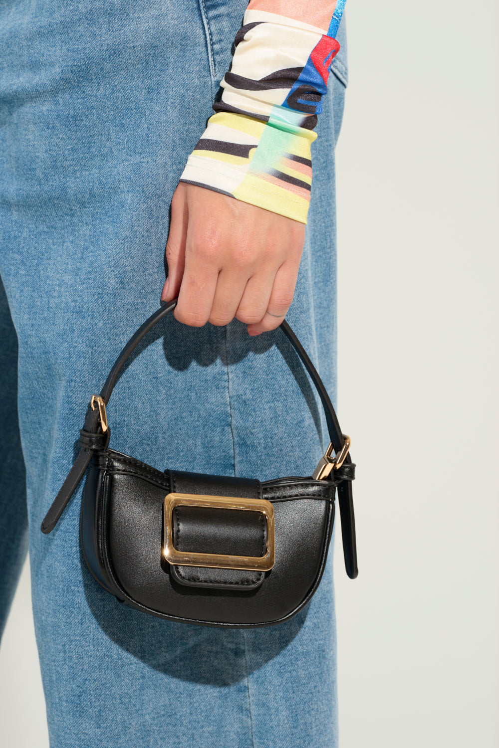 A person holding a QUIET LUXURY MINI PURSE featuring a faux leather finish and an oversized gold buckle, paired with blue jeans and a vibrant, striped long-sleeve top. 0