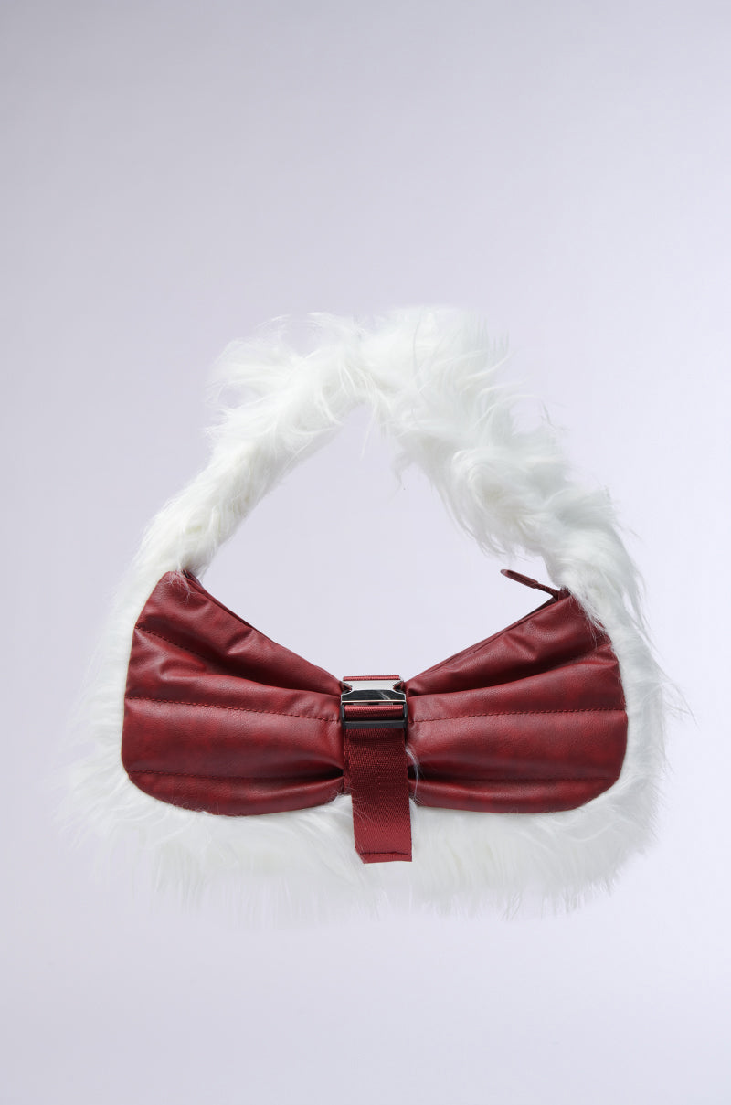 The OVERZEALOUS FUR TRIM PURSE from the AKIRA Label, showcasing a striking red color with a large bow design and elegant white faux fur trim, sits beautifully against a plain light background. Its charm is further enhanced by a central buckle and curved handle.