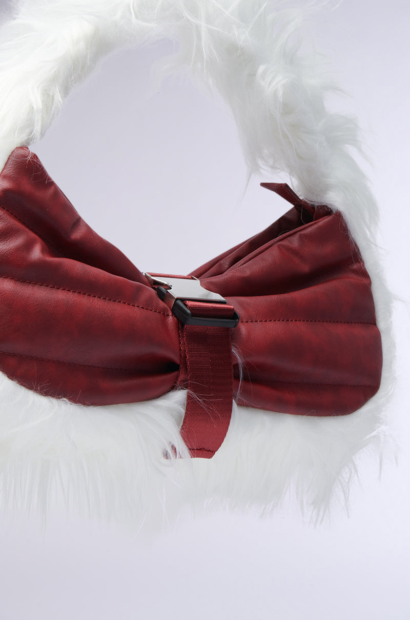 The OVERZEALOUS FUR TRIM PURSE from the AKIRA Label showcases a stylish design with a white faux fur trim and a red faux leather bow. It features a shiny metal buckle in the center, adding to its unique and fashionable appearance.