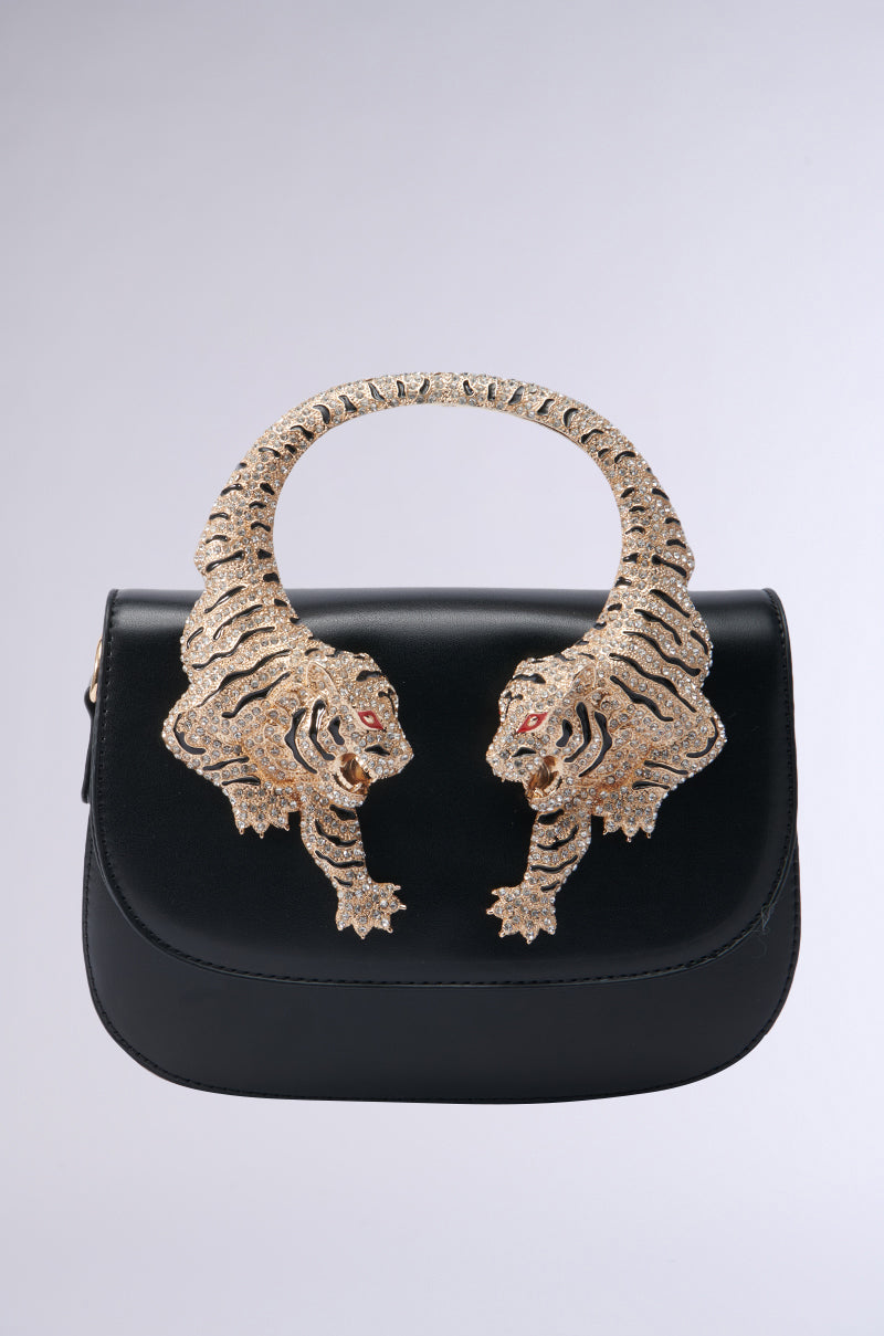 Introducing the EYE ON THE PRIZE LUXE BAG, a stylish black crossbody handbag that boasts an elegant design with a striking gold handle shaped like two tigers facing each other. The tigers’ red eyes and textured bodies provide an exquisite contrast against the light gray faux leather background.