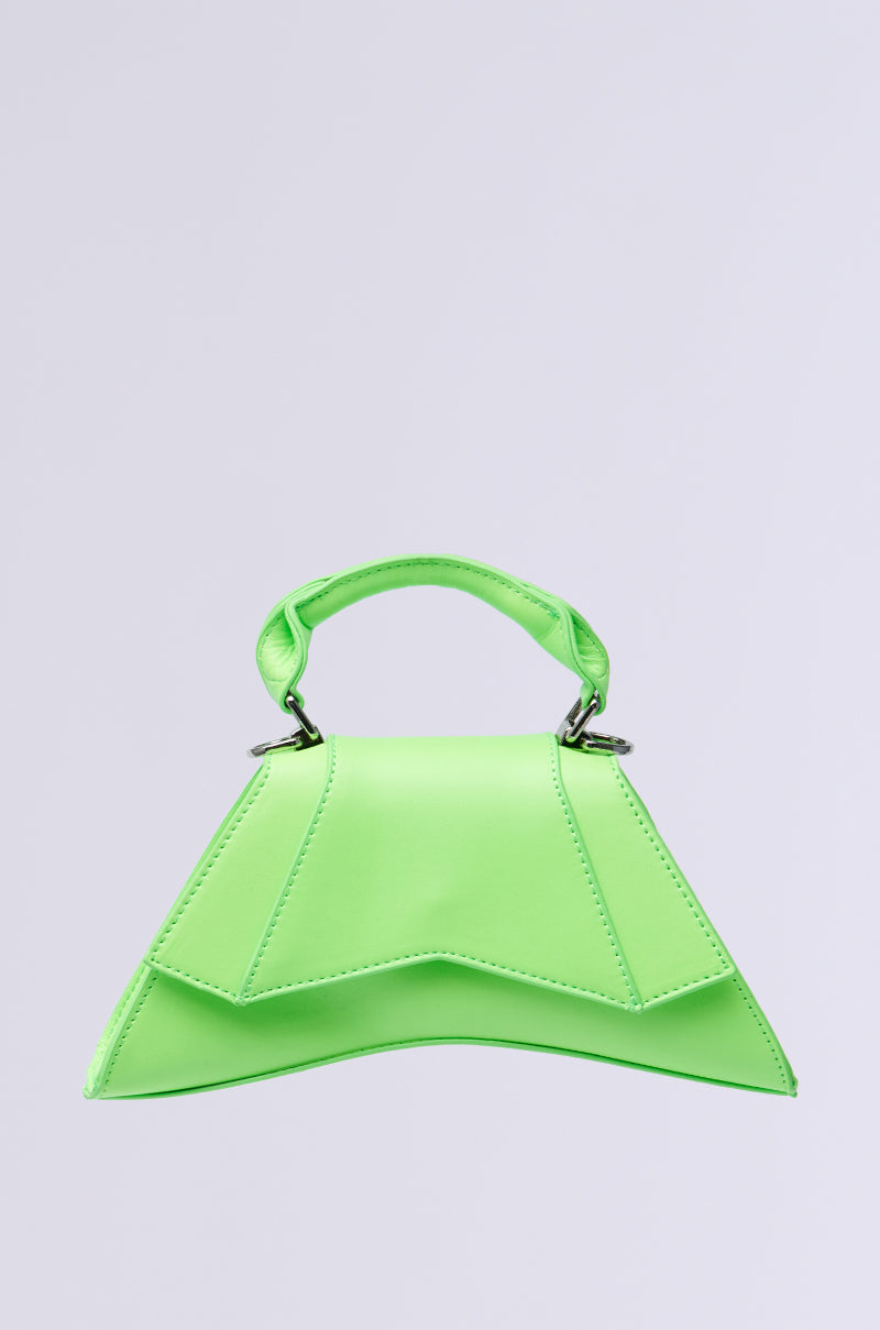 Against a gray background, the SO BRATTTTT MINI PURSE stands out with its vibrant green color, geometrically structured design, and hourglass silhouette. This handbag features a top handle and unique angular shape, giving it a modern and stylish appearance.