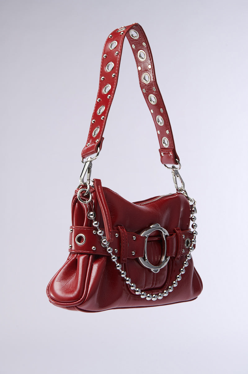 The RED HOT PU BAG is a structured handbag with a wide grommet-studded strap, large silver buckle, and ball chain. Its soft, gathered silhouette and shiny faux leather finish add bold style to any outfit.