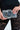 A person is holding the TAKE MY PIC PURSE, a faux leather handbag designed to look like a camera with lens and shutter button details, while wearing black lace pants.