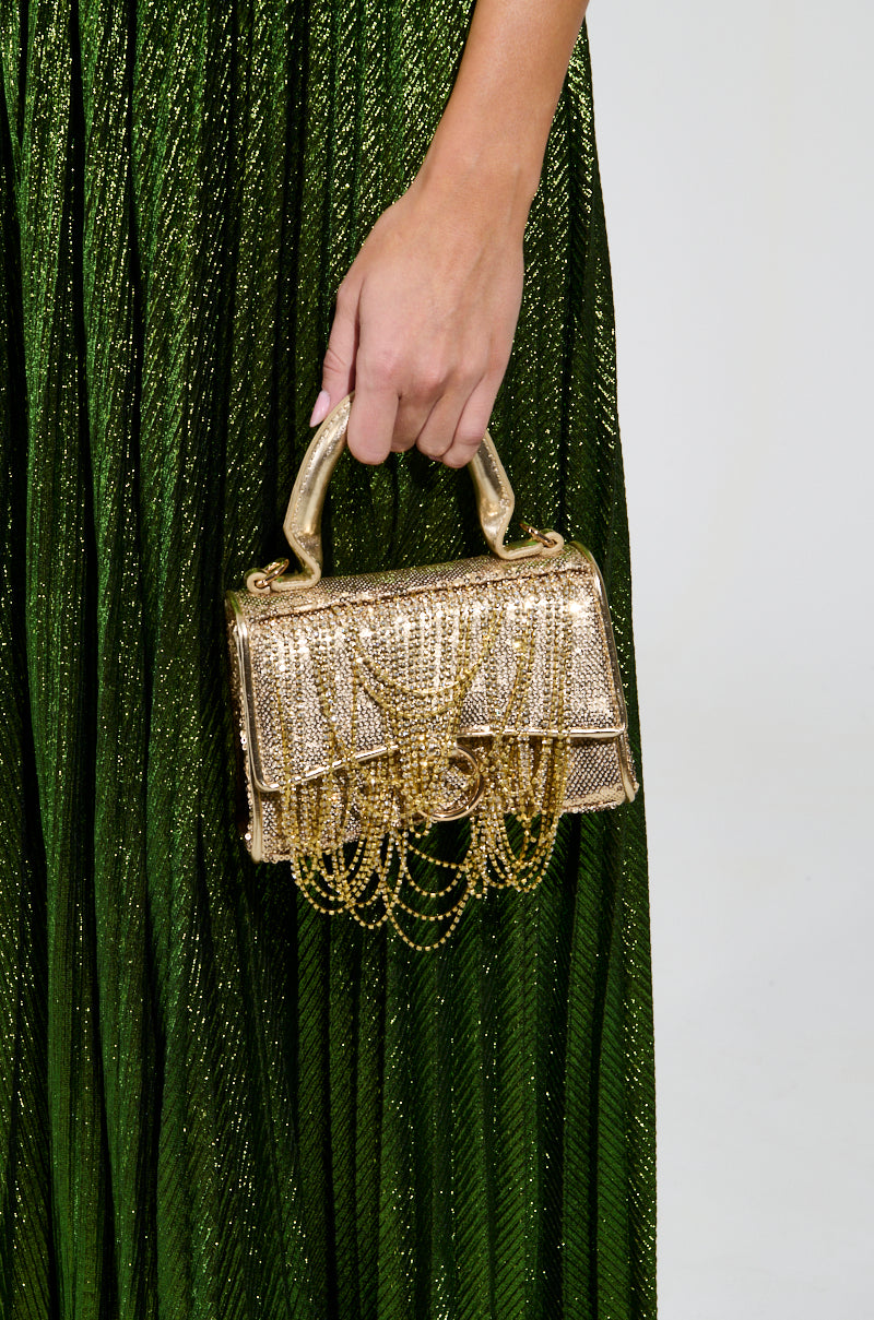 A person holds the KEEP DREAMING BLING PURSE, a small, ornate evening bag adorned with shimmering rhinestone fringe. The bag beautifully complements the pleated, metallic green skirt they are wearing.