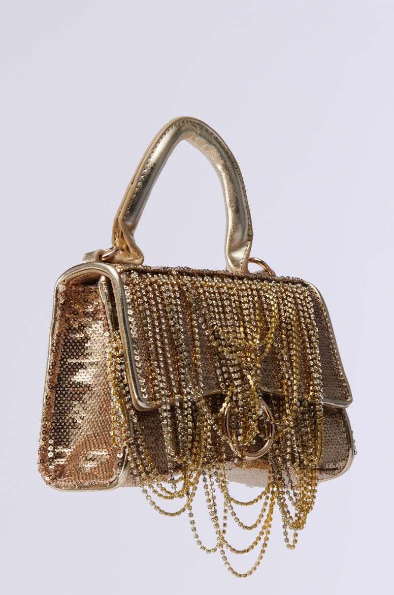 The KEEP DREAMING BLING PURSE is a glamorous gold evening bag adorned with shimmering sequins and rhinestone fringe, featuring a metallic handle and a prominent clasp that enhance its luxurious appearance against a light background.