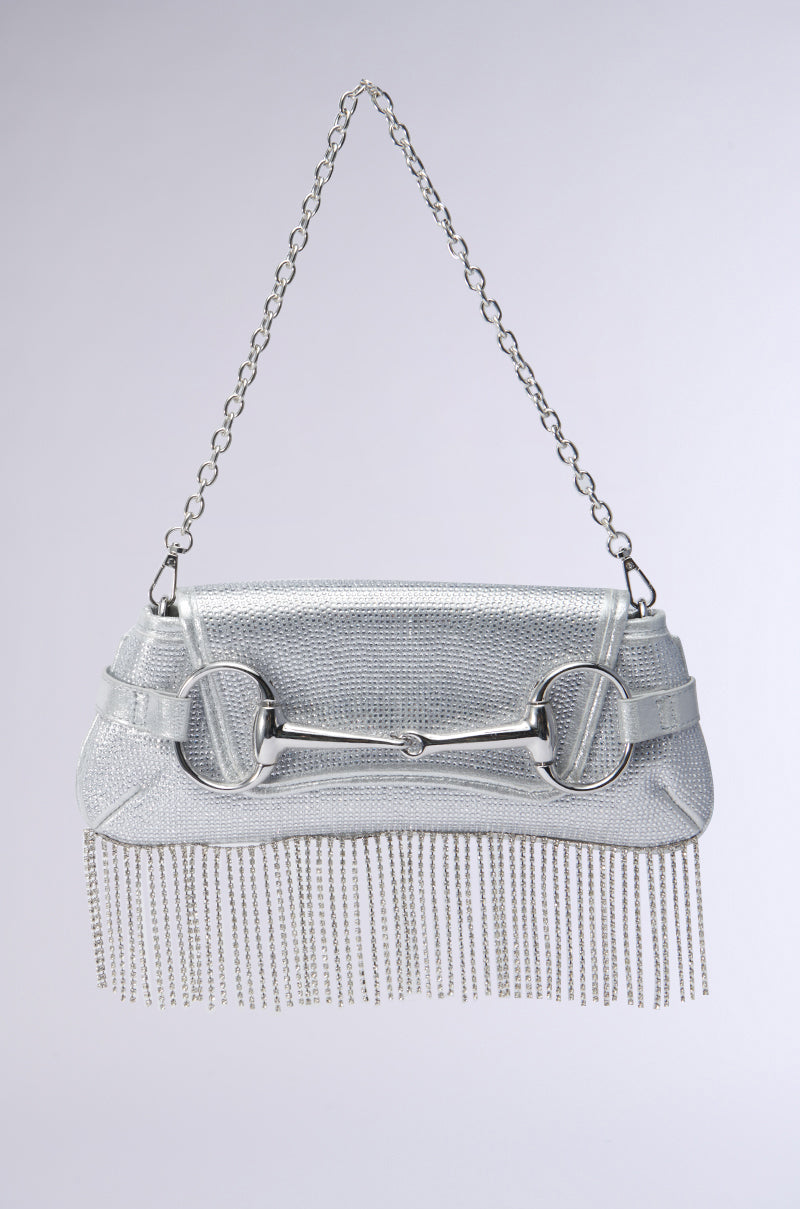 The FLY ME OUT BLING PURSE, a silver handbag with a chain strap and rhinestone fringe detailing, hangs against a light background. It showcases a large metal buckle decoration on the front, adding the perfect touch of bling.