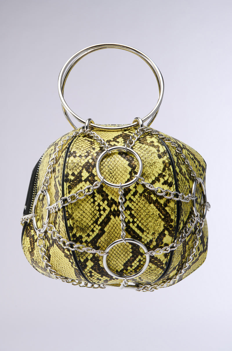 The MY STANDARD SNAKE BAG is a vegan leather handbag with a vibrant yellow snakeskin pattern and black accents. It features silver chains with circular metal rings, metallic chain details, and a unique handle of two intertwined metal hoops. 0