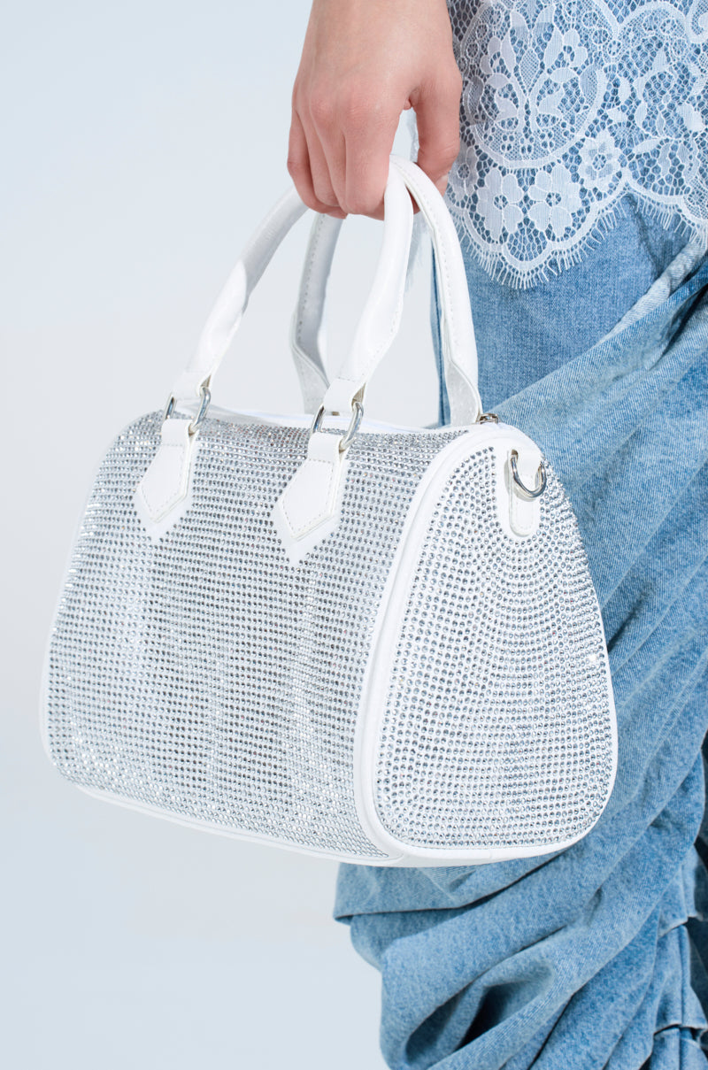 A person holds the BLING BOWLER BAG, featuring a sparkling white design with a silver rhinestone-like texture and a detachable shoulder strap. They wear blue draped pants and a white lace top against a light blue background. 1