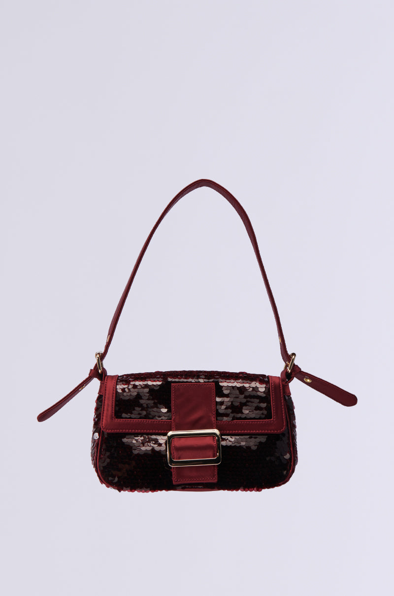 The MATCH ME SEQUIN PURSE is a chic festive baguette handbag with a dark red strap, shimmering sequin design, and a large rectangular faux gold buckle on the flap, providing a sleek minimalist look against a plain light gray background.