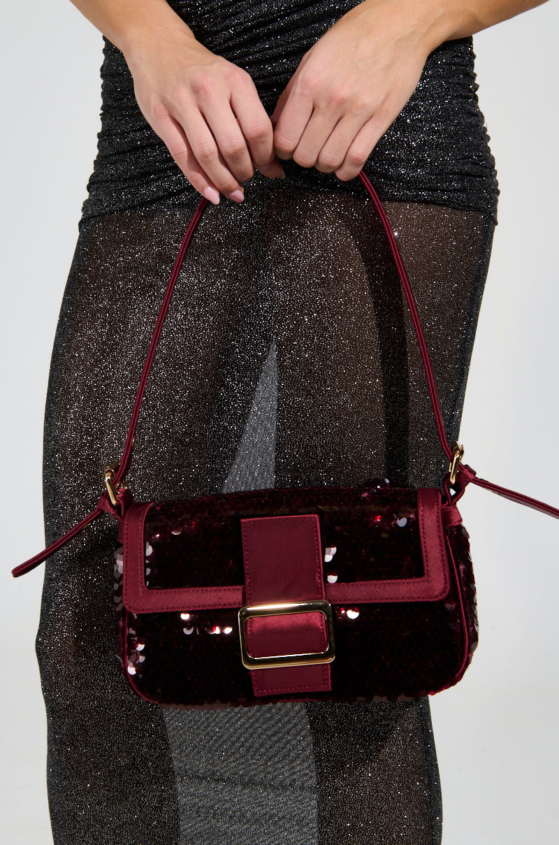 A person in a sparkly, sheer black dress holds the MATCH ME SEQUIN PURSE with faux gold hardware and a metallic buckle. Its shoulder strap and rich burgundy color contrast beautifully with the dress's glittery texture.