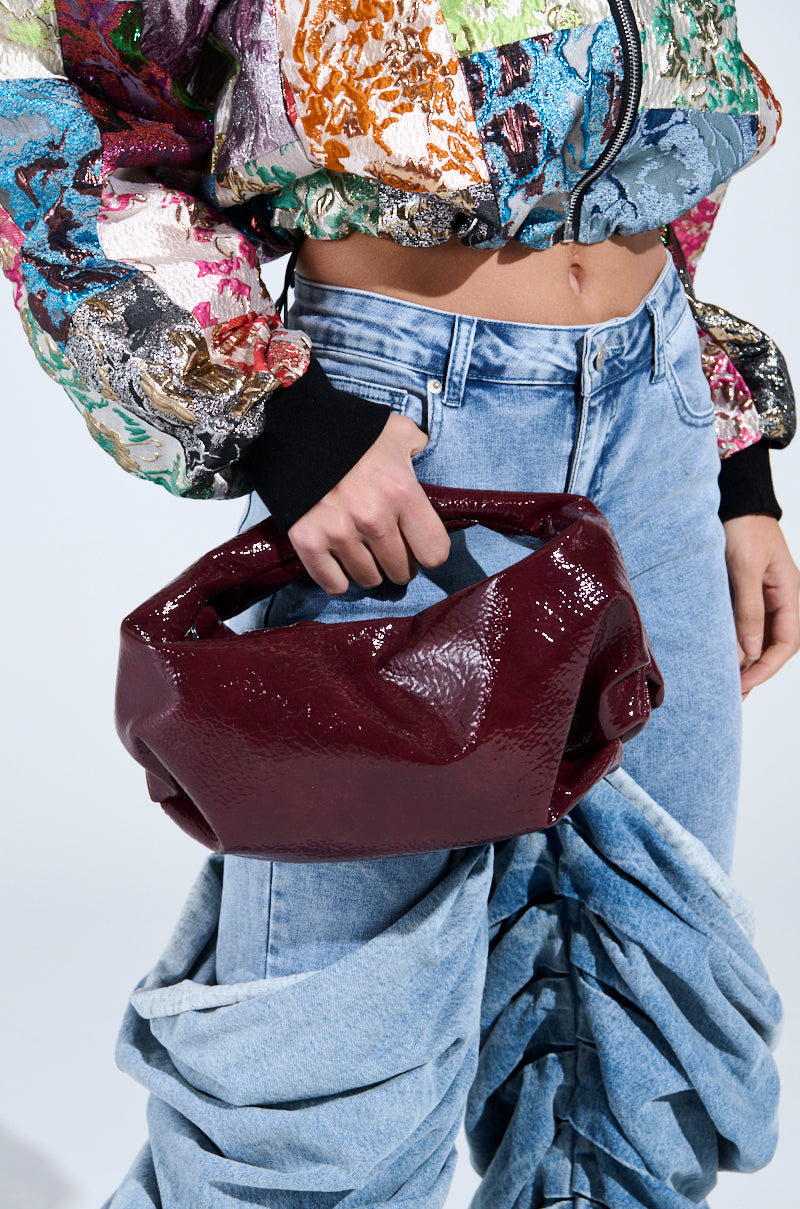 A person in a colorful textured jacket and high-waisted, ruched blue jeans holds the chic "Leather Lovin' PU Purse in Burgundy." The cropped jacket reveals their midriff, and the jeans boast a unique sculpted look. 1