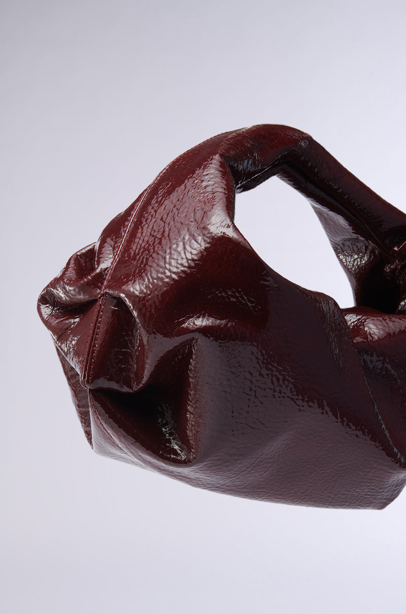 The LEATHER LOVIN' PU PURSE IN BURGUNDY is a stylish handbag, featuring a chic dark red faux leather with a shiny, crumpled finish and soft slouchy design. It has short handles and is beautifully displayed against a plain light gray background. 3