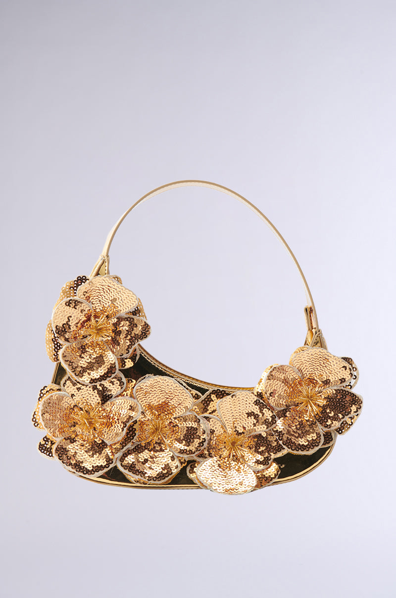 The FLORAL VIBEZ CHROME PURSE is a stylish handbag with a structured baguette silhouette and curved design, featuring large sequin-embellished floral appliques and chrome accents that sparkle against a soft, neutral gray background. A thin, elegant handle complements the intricate flower details.