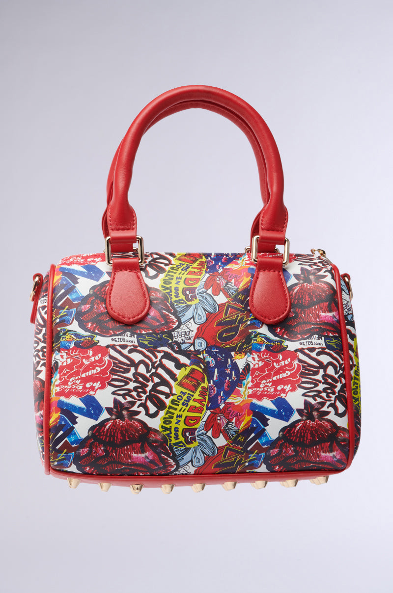 Explore the FLY ME OUT PURSE, a vibrant mini bowler bag with a graffiti-inspired pattern and comic book-style print. This faux leather handbag boasts red handles, gold hardware, and playful spikes at the bottom, showcasing graphic elements in red, blue, yellow, and black.