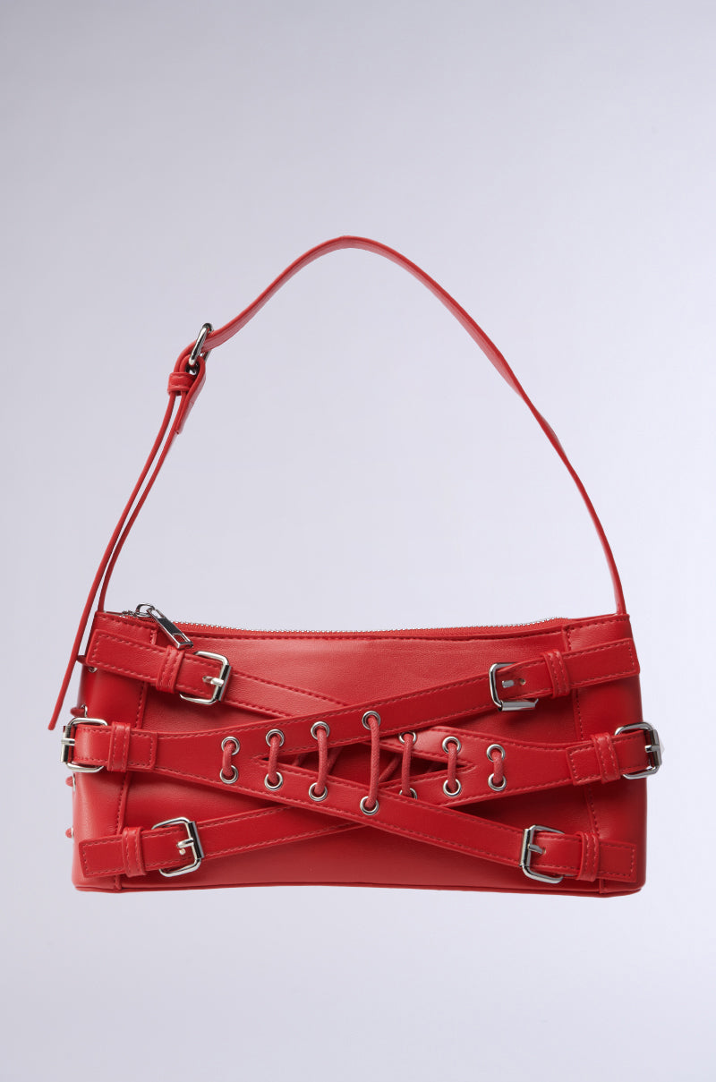 The CROSSED OUT SHOULDER PURSE is a red, vegan leather handbag with silver buckles and crisscrossing straps. It boasts a sleek design featuring a short shoulder strap and decorative metal eyelets on the front, set against a plain light gray background. 3