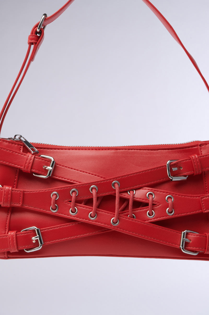 The CROSSED OUT SHOULDER PURSE is a red vegan leather handbag adorned with silver buckles and eyelets, featuring an intricate crisscross strap design on the front and a top-attached shoulder strap, set against a simple grey background. 5