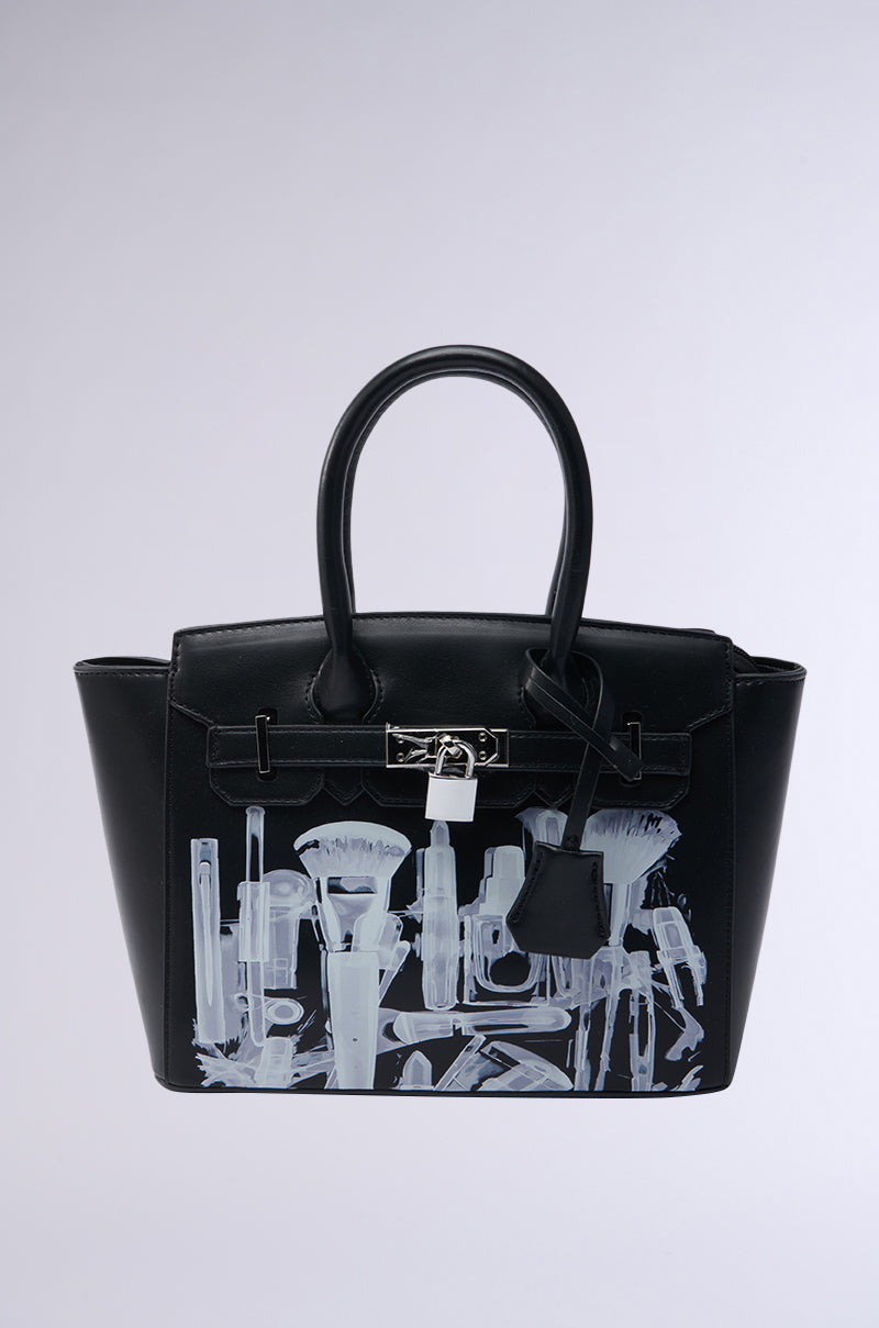 The SEE THRU ME TOTE is a black faux leather bag with silver hardware and top handles, showcasing a bold white X-ray print of makeup brushes and cosmetic tools on the front. 0