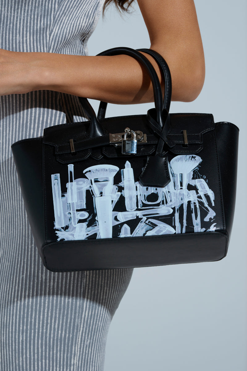 A person in a gray ribbed dress holds the SEE THRU ME TOTE, a faux leather bag with white X-ray graphics, lock detail, and double handles, set against a simple light gray background. 1