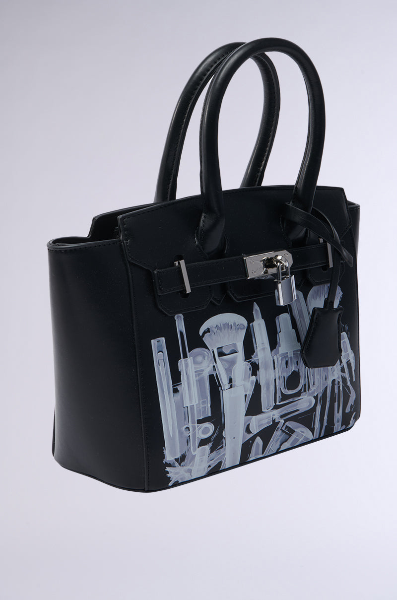 The SEE THRU ME TOTE is a black faux leather tote with double handles, silver hardware, and an abstract white and gray makeup brush-inspired design on the front, reflecting its signature X-ray graphic style. 2