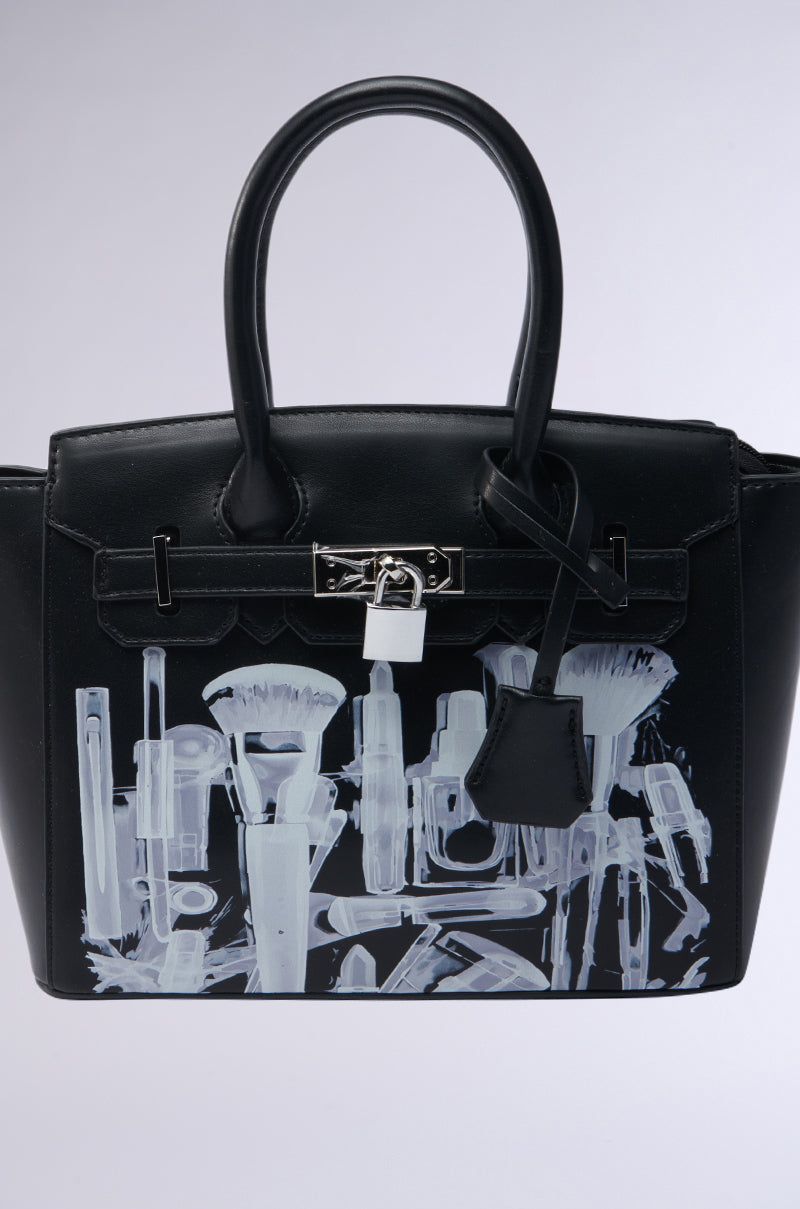 The SEE THRU ME TOTE is a black faux leather bag with silver hardware, two handles, a small attached tag, and white X-ray style makeup brush and cosmetic illustrations on the front. 3