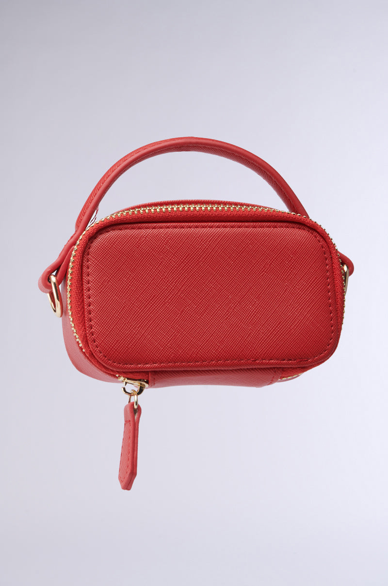The IN MILAN CROSSBODY CARD HOLDER is a chic, small red rectangular handbag with a textured surface, gold zipper, and strap. Made from vegan leather, it exudes elegance against a plain gray background.