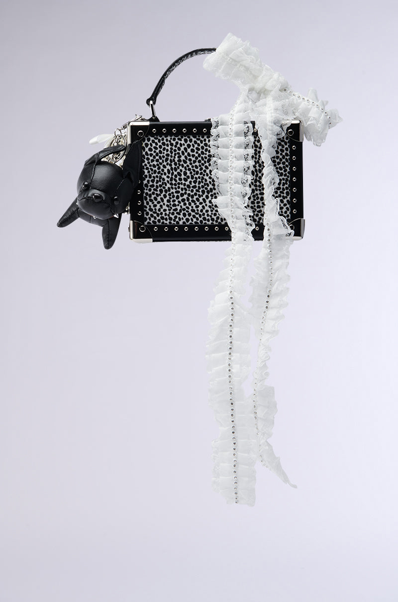 The KELYN JUNK BAG is a black rectangular faux leather bag with studded detailing, featuring a small black animal-shaped charm and a long ruffled white lace ribbon, shown on a plain light grey background.