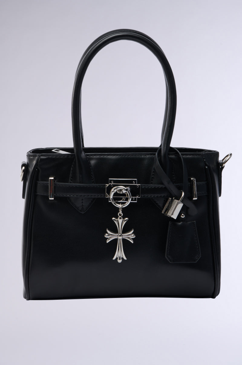 The CROSS MY HEART TOTE is a sophisticated black faux leather bag with silver hardware, a distinctive silver cross charm on the handle, and an elegant structured design. It includes a decorative lock, matching leather tag, and detachable shoulder strap. It is showcased on a plain gray background.