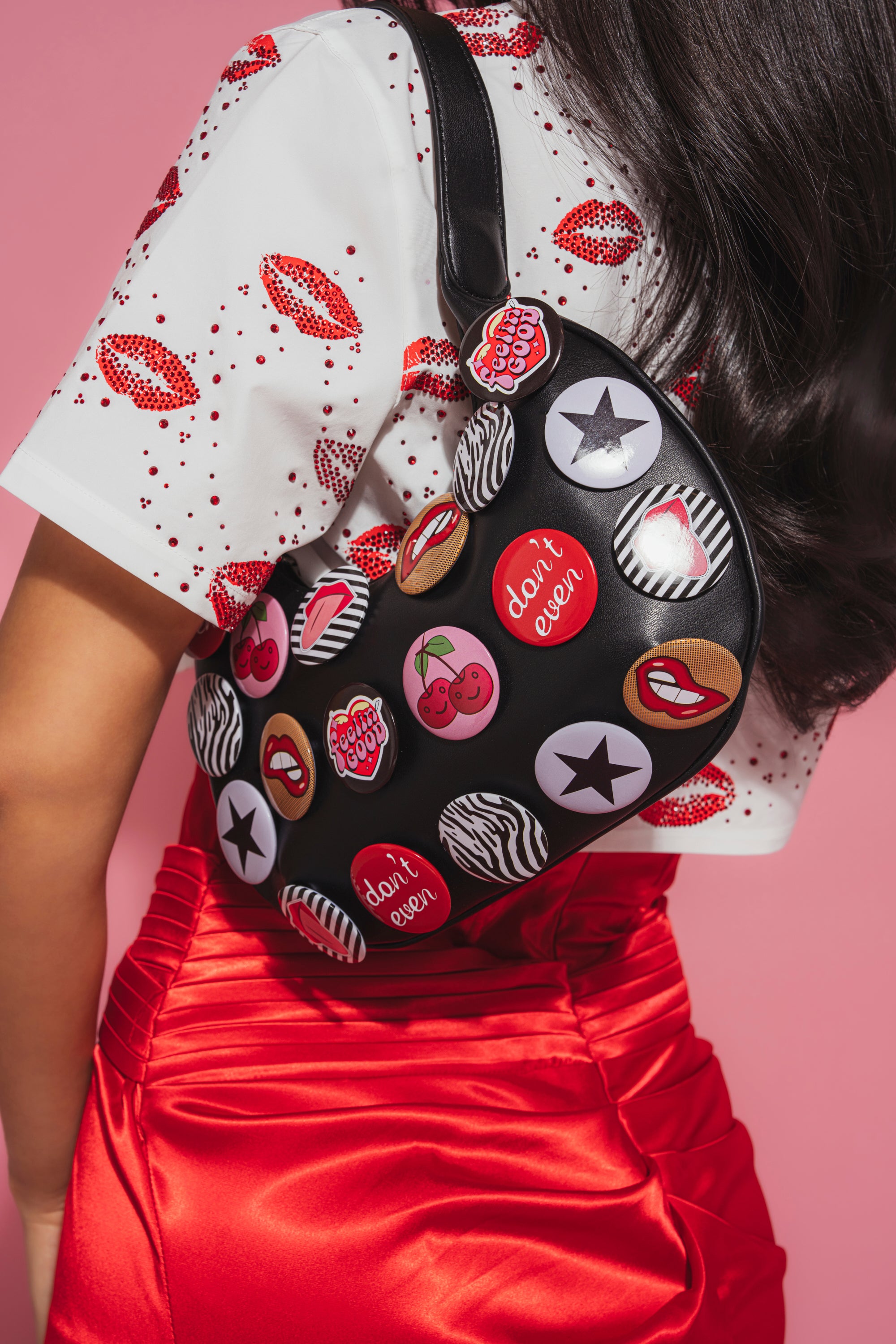 A person holds the LOVE ME BUTTON PURSE—a black faux-leather shoulder bag with colorful patches and “don’t even” graphic buttons—while wearing a white shirt with red lips print and a shiny red skirt, all against a pink background.