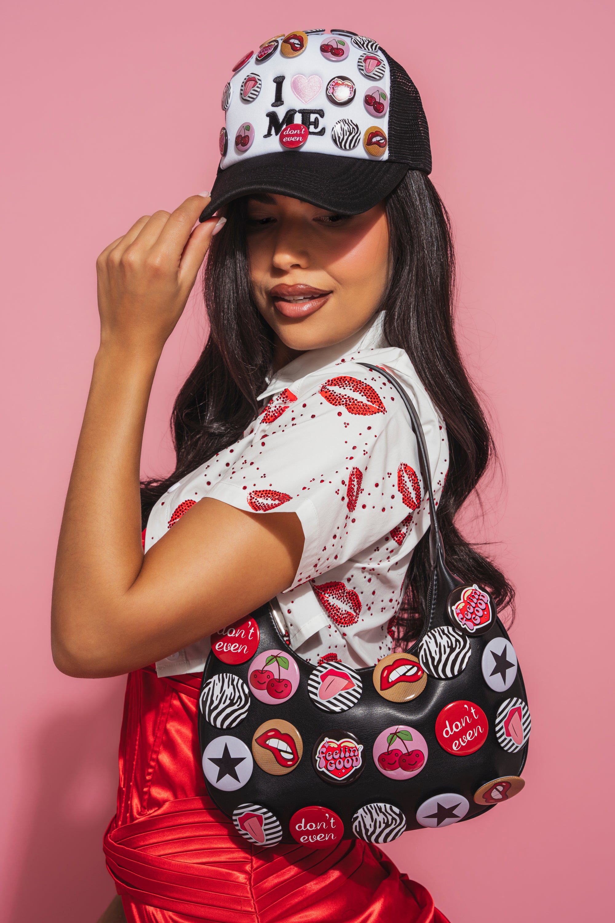 A woman in a white shirt, red lips, red satin dress, and a black-and-white cap poses against a pink background. She carries the LOVE ME BUTTON PURSE—a statement bag adorned with colorful badges and graphic patterns.