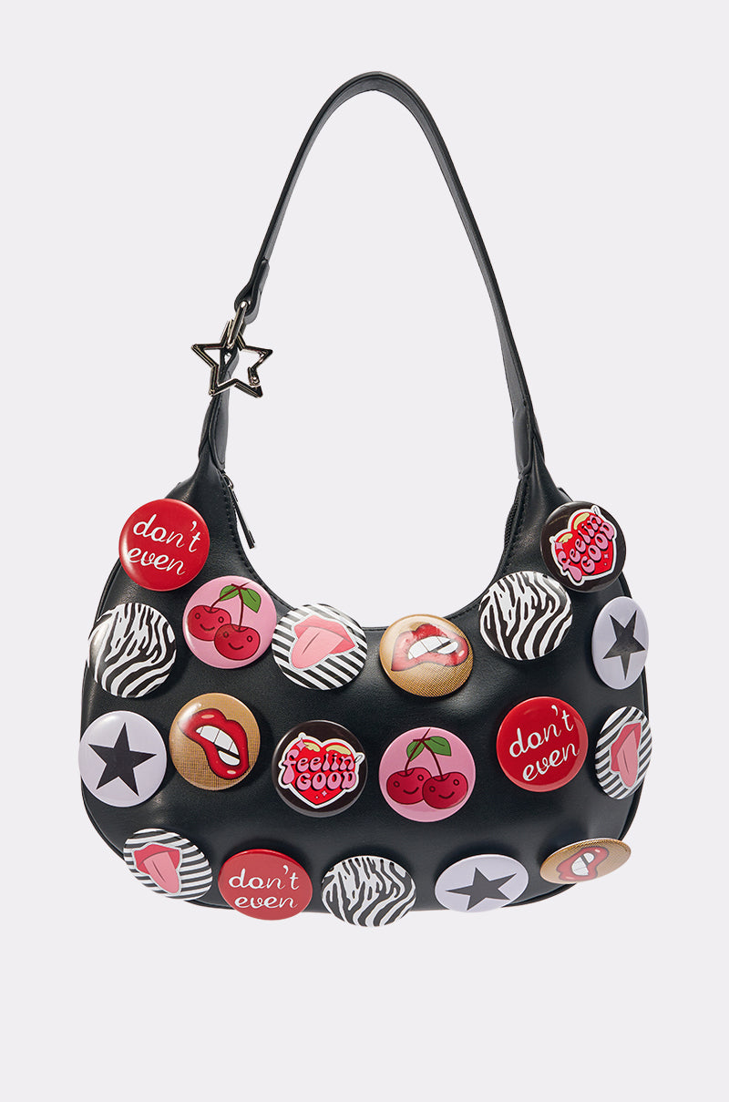 The LOVE ME BUTTON PURSE is a black crescent bag with a star-shaped handle and playful button embellishments—cherries, lips, stars, animal print, and phrases like "don’t even" and "feelin’ good"—for a bold statement look.