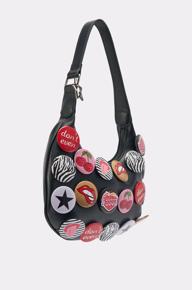 The LOVE ME BUTTON PURSE, a black faux-leather bag with vibrant button accents—phrases, cherries, lips, a star, and zebra stripes—stands out boldly against a simple light backdrop.