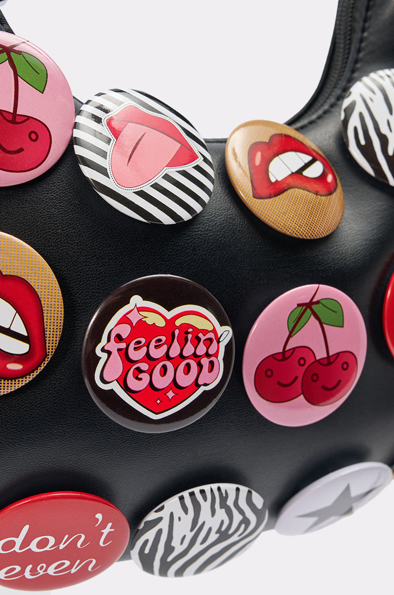Close-up of the LOVE ME BUTTON PURSE, a black faux-leather bag adorned with vibrant button accents—lips, cherries, zebra prints, and a "feelin' GOOD" pin—making it the ideal playful statement accessory.