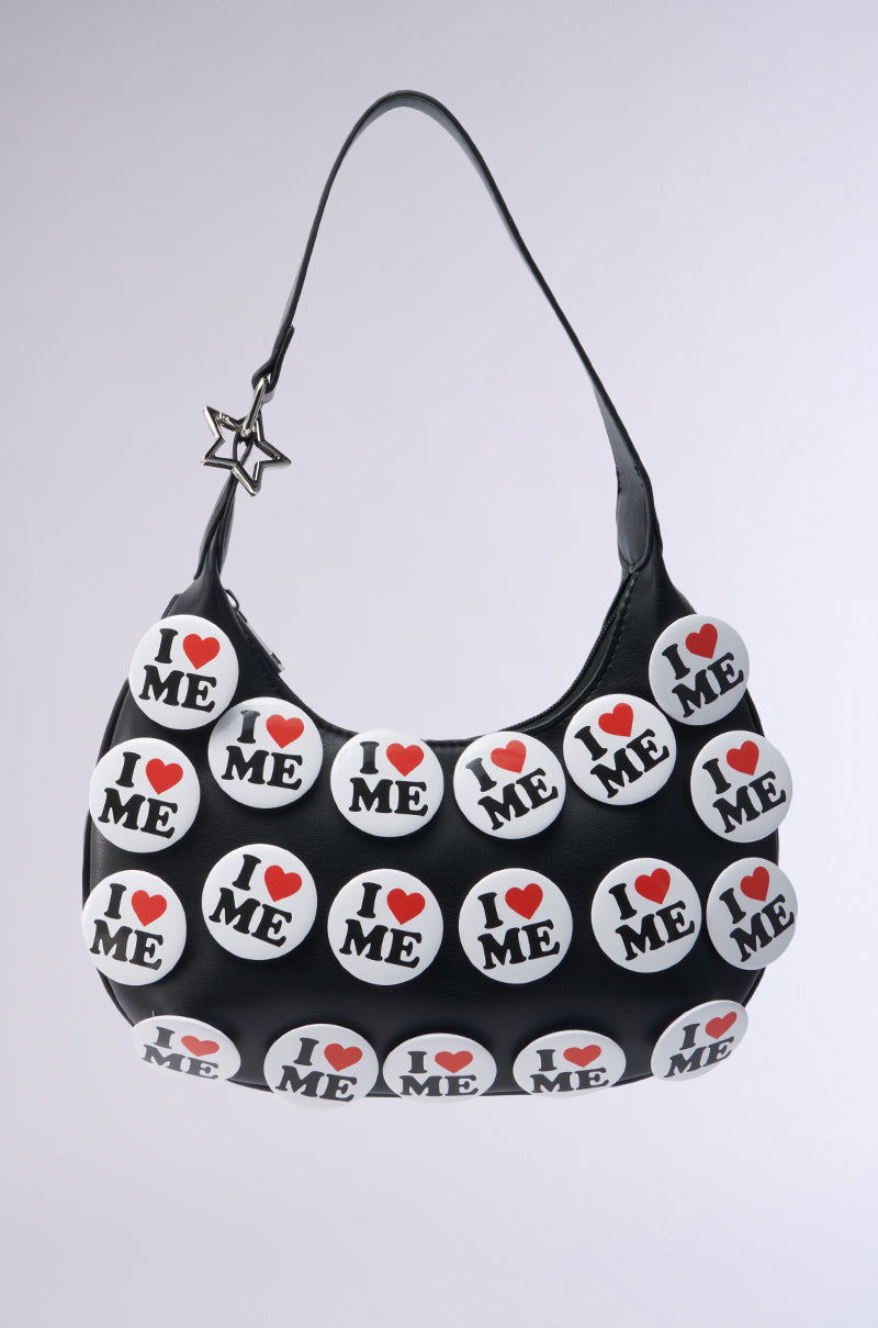 The "I REALLY DO PIN PURSE" is a black faux leather handbag adorned with bold "I ♥ ME" buttons in black and red. A star-shaped charm hanging from the handle enhances its sassy fashion appeal, all set against a plain white background.