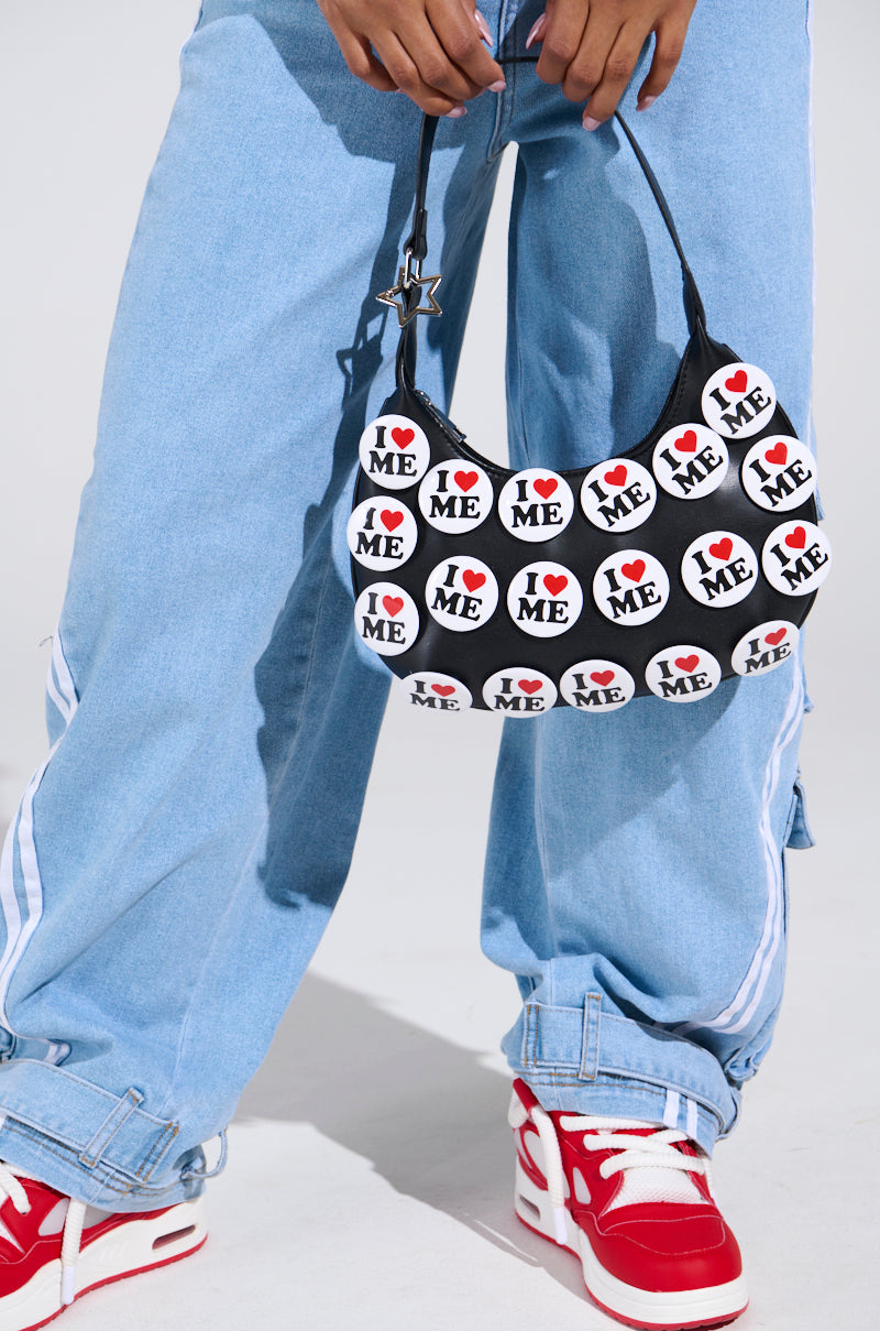 In light blue jeans and red sneakers, a person holds the I REALLY DO PIN PURSE: a sassy faux leather accessory featuring circular "I ❤️ ME" pins, a short strap, and clasp. This purse stands out against the plain background, showcasing its casual and expressive style.